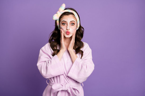 年轻女孩在明亮的紫色背景下用泡沫清洁肌肤，涂抹保湿霜并送出空气吻的照片肖像图片下载