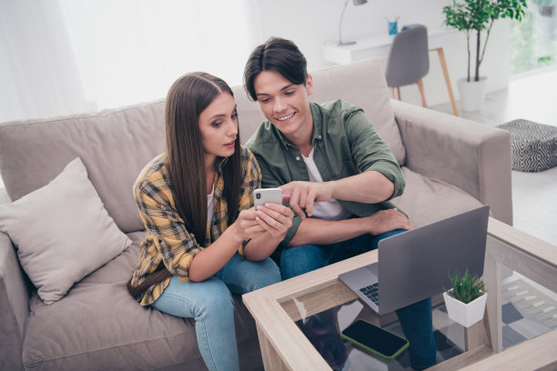一对年轻的情侣穿着休闲服，坐在沙发上微笑着阅读现代设备，室内公寓的照片。图片下载