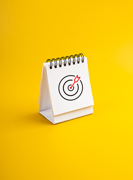 白色桌面日历页上的靶心图标，背景为黄色，竖版照片。商业目标与成功、日程安排、议程、行动计划概念，适合新年。图片下载