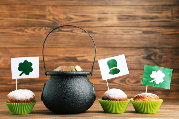 装满硬币和杯形蛋糕的罐子，上面插着旗帜和帽子、三叶草的图片，背景是棕色木纹。图片下载