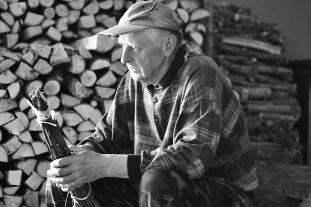 黑白照片中，一位年长的农民坐在堆放的柴火前，手中拿着一把手工制作的地刷。这个乡村场景反映了传统农村生活、工艺和简单的生活方式。图片下载