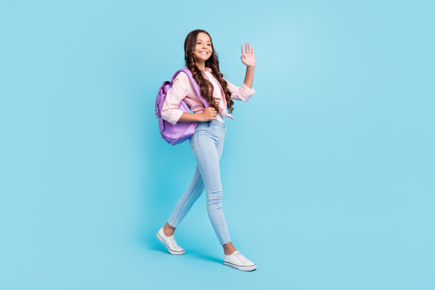 年轻女孩微笑着，心情愉快，走向学校，在蓝色背景下打招呼的全身照片图片下载