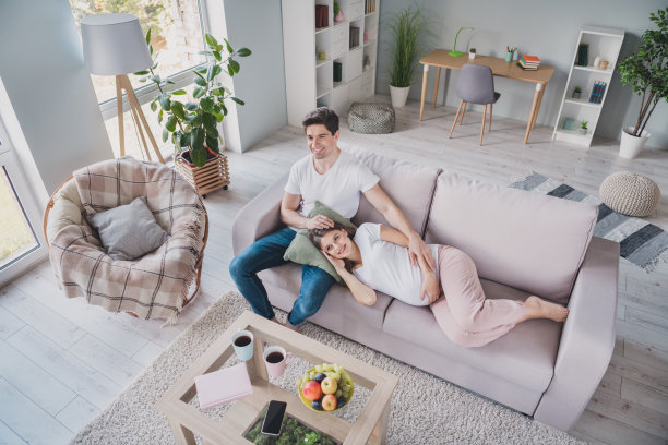年轻家庭在家中沙发上享受周末，坐着看电视，手放在肚子上，俯视全身的高角度照片图片下载