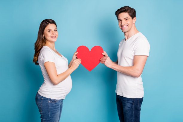 乐观的孕妇情侣穿着白色T恤和牛仔裤，手持纸质心形，背景为青色。图片下载
