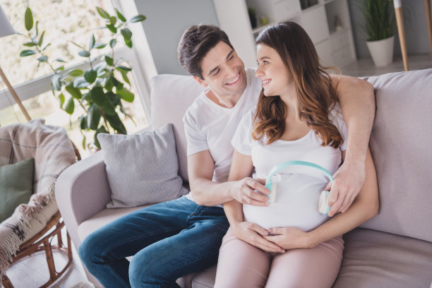 可爱的甜美家庭侧面照片，坐在沙发上抱着肚子，戴着耳机，室内等待宝宝。图片下载