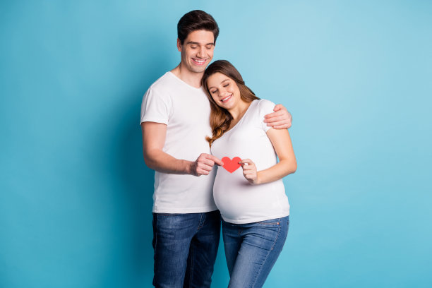 一对夫妻的照片肖像，丈夫怀抱着怀孕的妻子，手中握着红色心形物体，背景为鲜艳的蓝色。图片下载