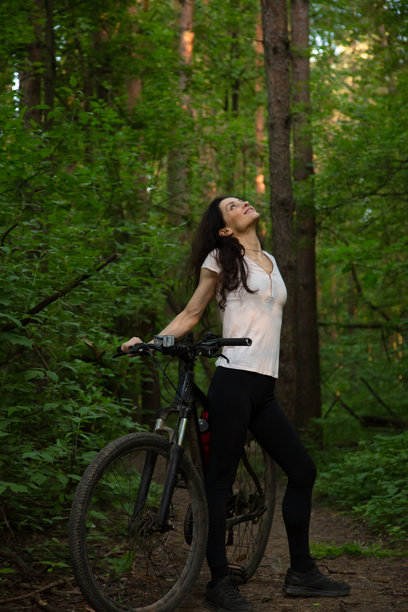 美丽的运动女性在夏季森林的绿树林间骑自行车，微笑着仰望。强壮的身体，腿部线条优美，女孩在户外骑行。女性在公园里骑着运动山地车。图片下载