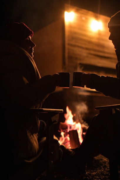这张照片展示了两个人在夜晚的篝火旁坐在椅子上。他们分享着杯子，杯中冒着热气。火焰的温暖光芒营造出一种舒适的氛围。图片下载