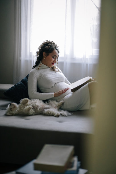 年轻的孕妇在公寓的沙发上享受她的日子，正在阅读一本书，她的宠物狗陪伴着她。图片下载