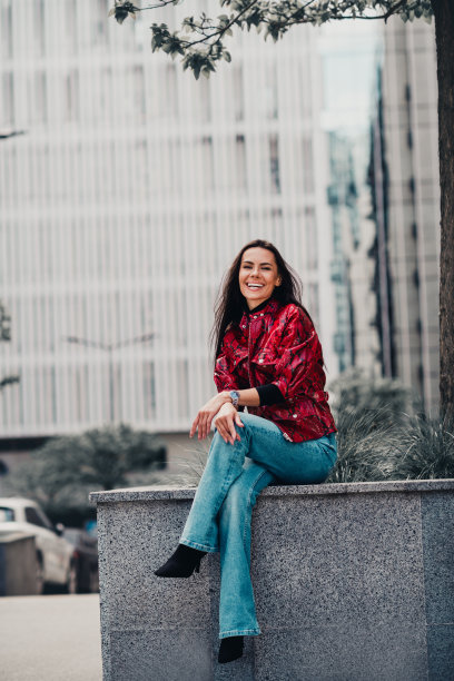 时尚的年轻女性，面带灿烂的微笑，穿着休闲的红色印花夹克和牛仔裤，背景为城市环境，竖版照片图片下载