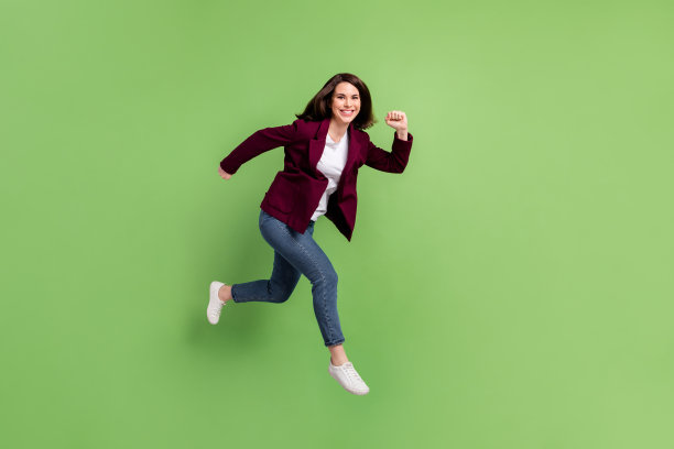 乐观的棕发女士穿着开衫、牛仔裤和运动鞋，跳跃着，背景为绿色，展示全身侧面照片。图片下载