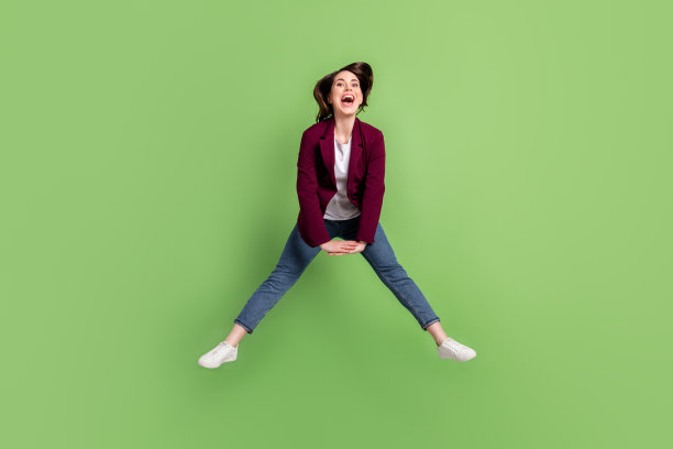 乐观的棕发女士穿着开衫、牛仔裤和运动鞋在绿色背景上跳跃的全身照片图片下载