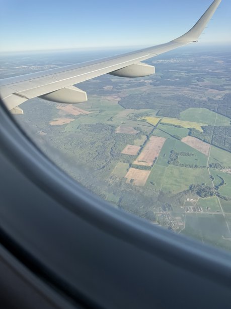 从飞机窗户拍摄的照片，展示了机翼和下面的风景，包括田野、森林和远处的城市，天空晴朗。图片下载