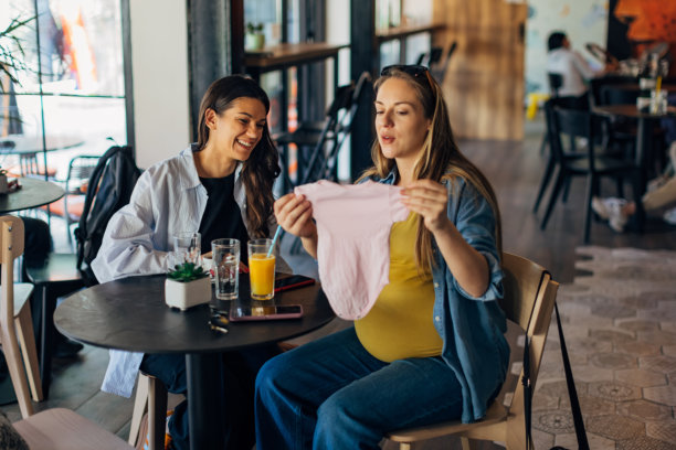 微笑的孕妇与她的好朋友一起看婴儿衣物。女性朋友正在给孕妇送一份新生儿的礼物。图片下载