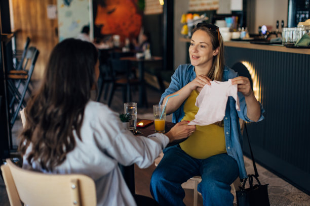 一位无忧无虑的孕妇与她的好朋友一起看婴儿衣物。女性朋友为孕妇送上了新生儿的礼物。图片下载