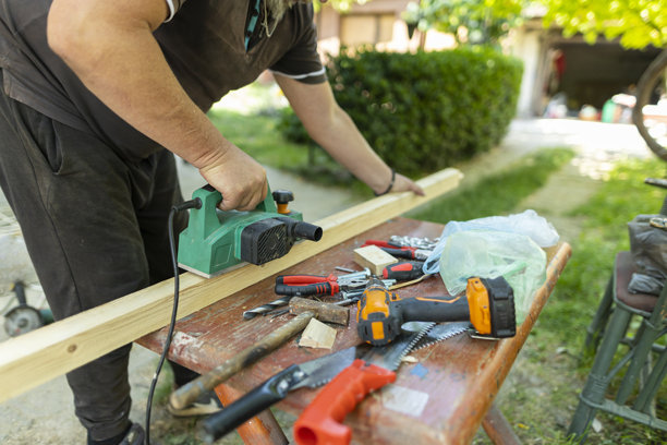 老年男子在户外使用电动刨子修整木板，周围摆放着各种手工具。家居改善和木工概念。特写照片图片下载