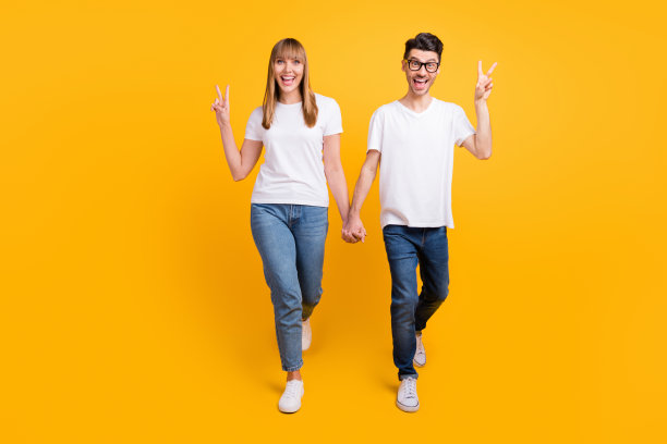 穿着T恤的可爱漂亮的人们在黄色背景下走路，手臂相互搂着，做出V字手势的全身照片图片下载