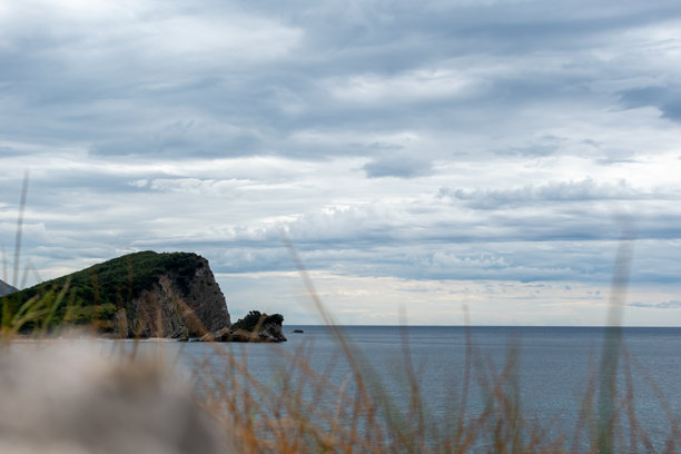 风景照片捕捉到一幅戏剧性的景象：一座崎岖的、绿顶悬崖突入平静的深蓝色海洋，广阔的多云天空下，前景的高草模糊不清。图片下载