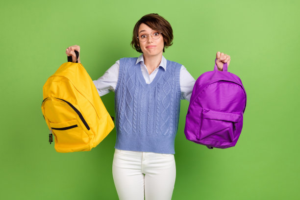 戴眼镜的不确定学生在鲜艳绿色背景下比较两个背包的照片肖像图片下载