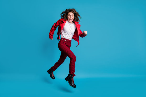 穿着红色夹克、裤子和靴子的欢呼女孩在蓝色背景上跳跃奔跑的全身侧面照片图片下载