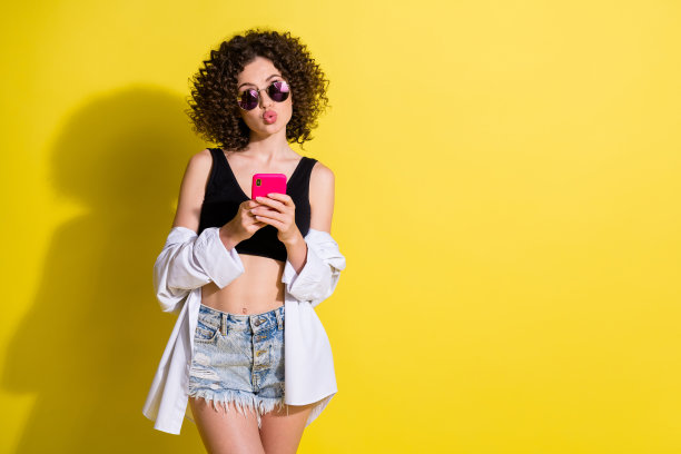 卷发女孩在明亮的黄色背景下用智能手机浏览互联网，送出空气吻的照片肖像，背景留有空白。图片下载