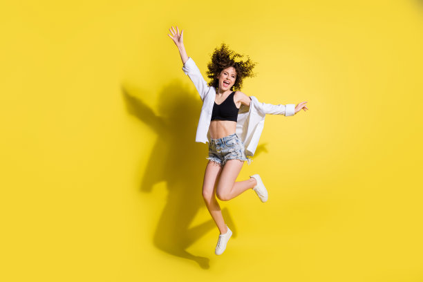 充满乐观的可爱女孩穿着黑色上衣、短裤和运动鞋在黄色背景上跳跃的全身照片图片下载