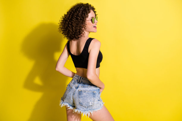 背影照片，女性穿着黑色上衣和牛仔短裤，戴着太阳镜，微笑，背景色彩鲜艳。图片下载