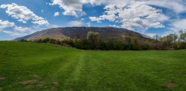 壮丽的山间绿草甸全景。草甸上方是蔚蓝的天空，点缀着白色的绵云。背景中可见一座巨大的山脉。春季或夏季的自然照片。图片下载