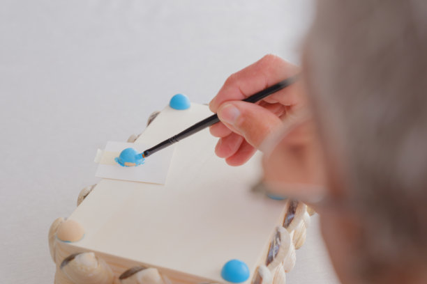 老年男子从肩后视角为手工贝壳首饰盒的腿部涂上蓝色。图片下载