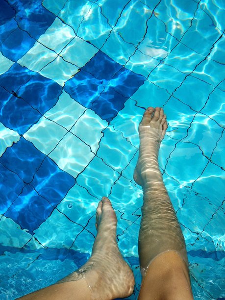 这张图片捕捉到一个人放松的时刻，双脚浸泡在游泳池中，池底是蓝白相间的方格瓷砖图案。图片下载
