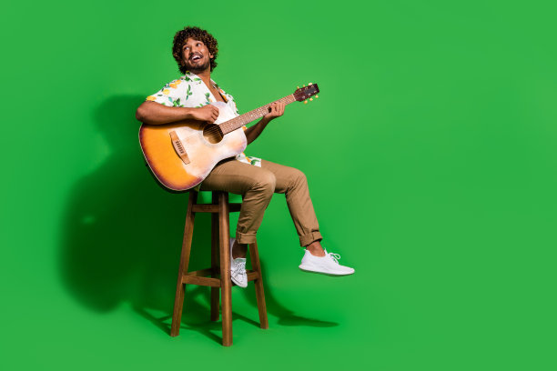 漂亮的年轻男性的全尺寸照片，坐椅子，吉他，看起来空的空间，穿时髦的柑橘印花服装孤立的绿色背景图片下载