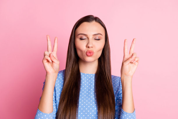 女友微笑着做出V字手势，送出空气吻，穿着蓝色波点服装，背景为粉色柔和色调的照片肖像。图片下载