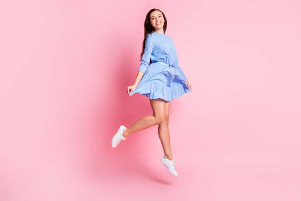 穿着蓝色波点裙的美丽女孩在粉色背景上高高跳起，面带微笑，展现全身的照片。图片下载