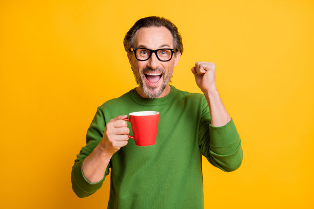 戴眼镜的男子在鲜艳的黄色背景下，像赢家一样手势庆祝，喝着咖啡，享受休息时光的照片肖像。图片下载