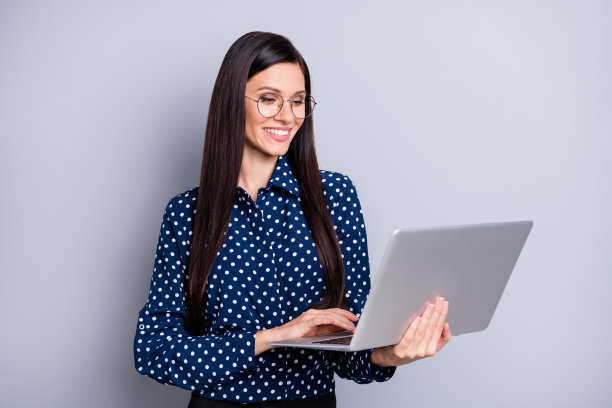 乐观的棕发女孩穿着蓝色衬衫和裤子，戴着眼镜，坐在灰色背景前使用笔记本电脑的照片图片下载