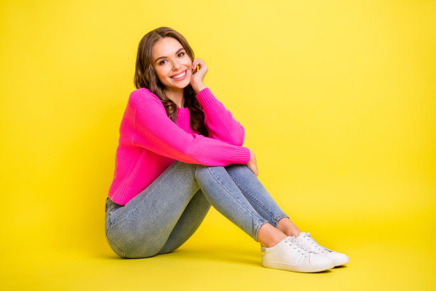 年轻漂亮女孩的全身侧面照片，开心地微笑，坐在地板上，背景为黄色。图片下载