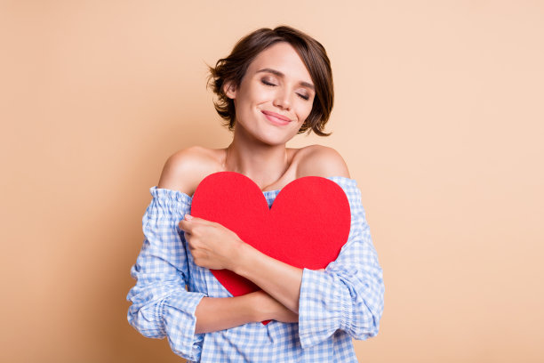 放松的女性双手抱着一张大红心卡片的照片肖像，背景为柔和的米色。图片下载