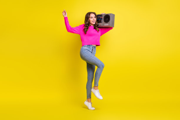 可爱的波浪卷发女性穿着粉色毛衣，肩上扛着音响在黄色背景下跳舞的全身照片图片下载