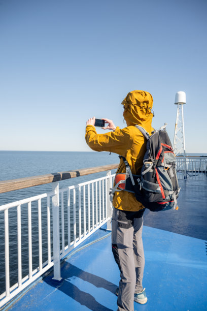 穿着夹克的旅行者在海上渡轮甲板上拍摄智能手机照片的背影图片下载