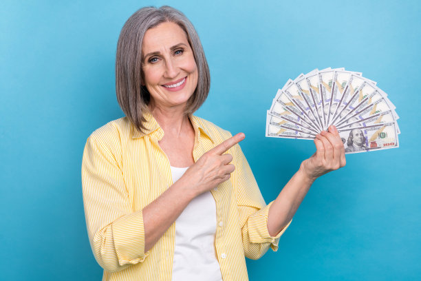 照片中开朗漂亮的女士穿着黄色衬衫，手指着现金扇孤立的蓝色背景图片下载