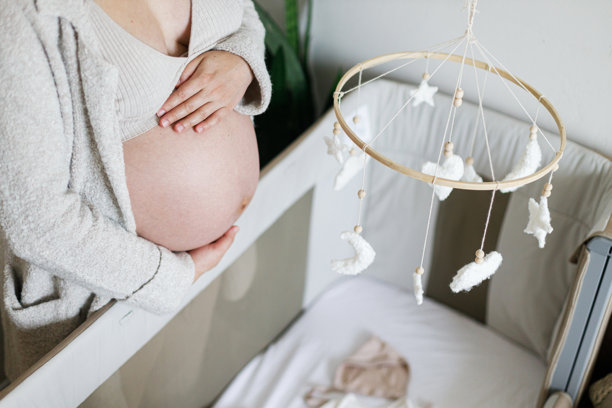 孕妇抱着肚子，站在婴儿床旁，床上有摇铃、超声波照片、衣物和玩具。怀孕。婴儿房准备。图片下载