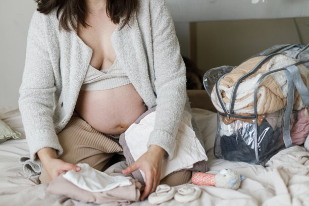 怀孕。为宝宝准备和购物。孕妇坐在舒适的床上，手里拿着婴儿衣物，正在打包分娩必需品的医院包。分娩日清单。图片下载