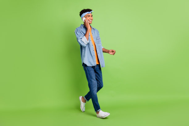 乐观的棕发男孩穿着乐队T恤、牛仔裤和运动鞋，站在绿色背景前，正在打电话的全身侧面照片。图片下载