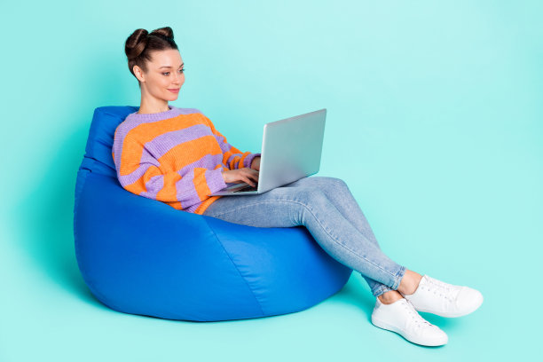 乐观的棕发女士坐着，穿着毛衣，手持笔记本电脑，背景为青色，展现出全身侧面照片。图片下载