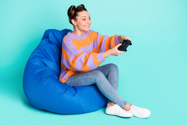 乐观的棕发女士穿着毛衣坐在PlayStation前，背景为青色，展示全身侧面照片。图片下载