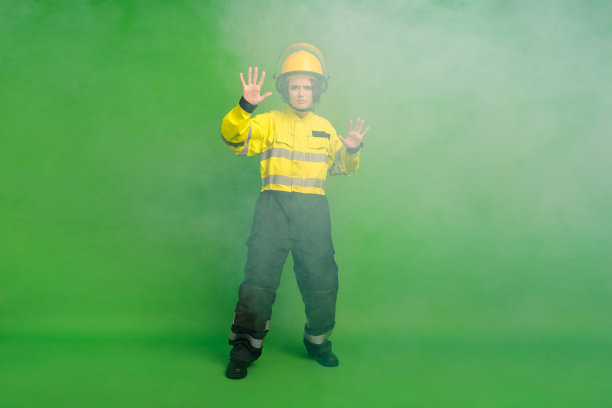 全身照片，充满自信的年轻女消防员，穿着工作服，戴着黄色安全帽，面临火焰和烟雾，背景为绿色。图片下载