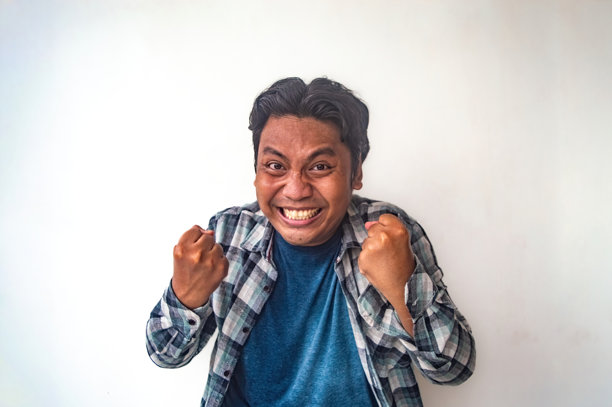 一名穿着格子衬衫和蓝色T恤的男子在简单的白色背景前握紧双拳，展示出成功或成就的手势。这张图片捕捉到了胜利和喜悦的瞬间。图片下载