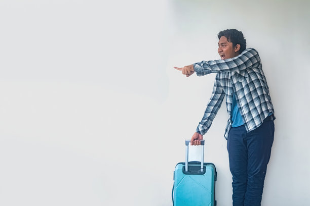 一名穿着格子衬衫和蓝色裤子的男子正拿着一个蓝色行李箱，面露惊讶的表情并指着前方。这张图片捕捉了旅行中惊讶或发现的瞬间。图片下载
