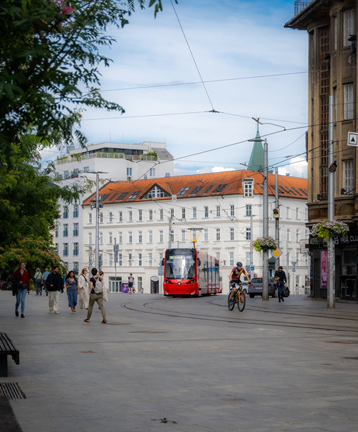垂直照片展示了一辆红色电车在布拉迪斯拉发市街道上，夏日里人们在步行和骑自行车。图片下载