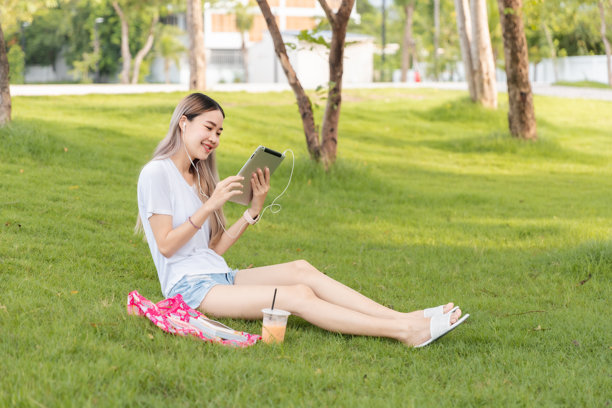 享受闲暇时光的快乐女性坐在公园的草地上，戴着耳机，玩着iPad，阳光明媚。空旷的空间。图片下载